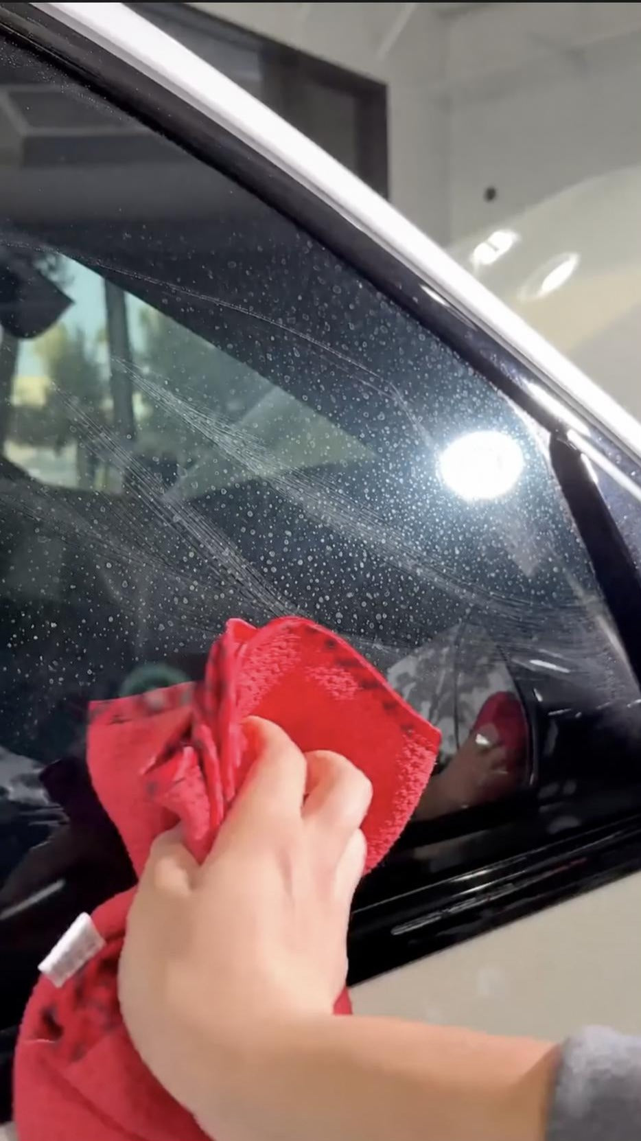Hand wiping a car window with a red microfibre cloth, with mineral spots still left on the glass